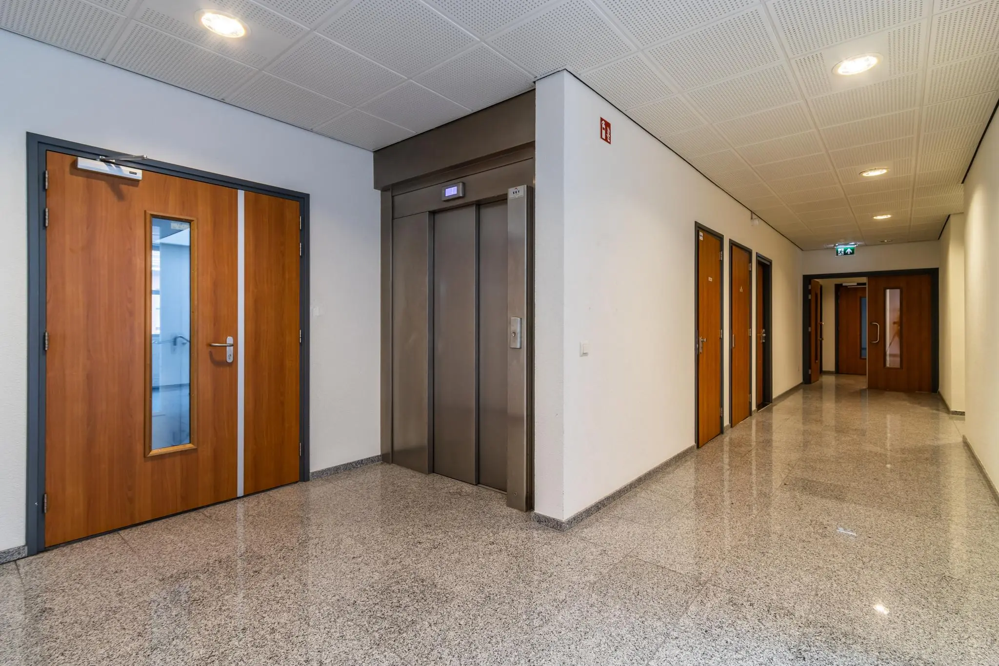 Hal met granieten vloer, houten deuren en een metalen lift in een kantoorgebouw aan de Lange Dreef.